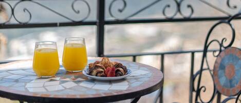 A round table with two glasses of orange juice and a plate of pastries on a balcony with a metal railing and a view of the mountains.