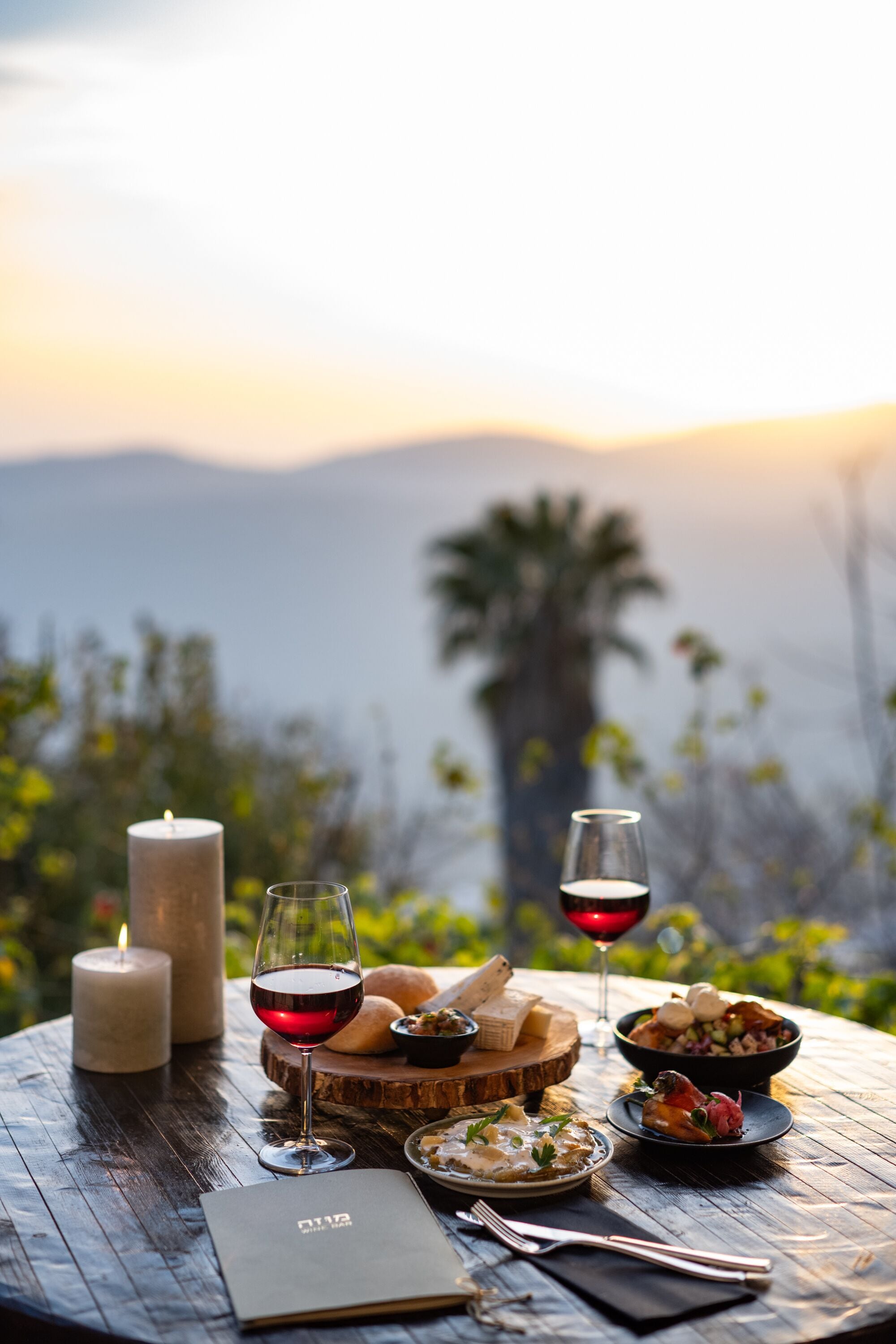 A table set with food and wine, candles, and a book, overlooking a mountainous landscape.