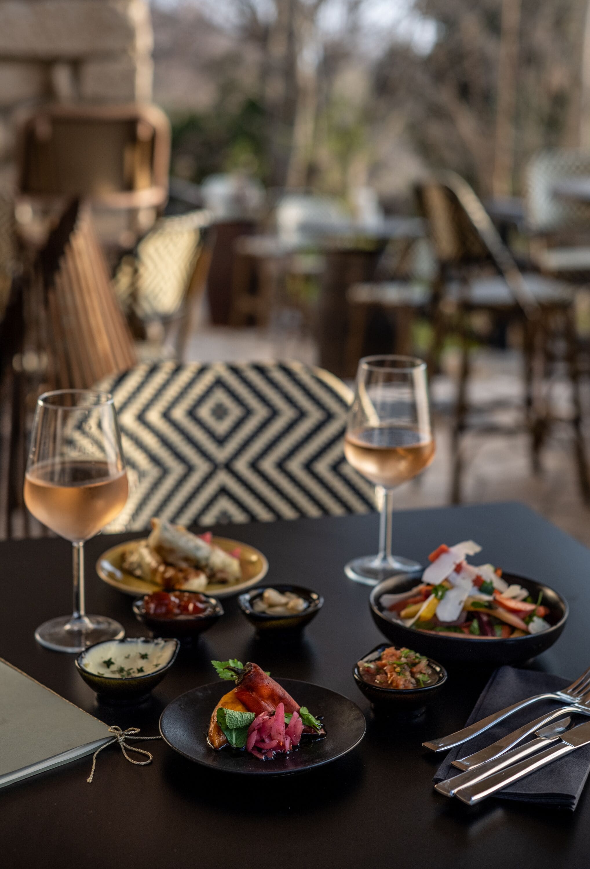 A dining table with various dishes and glasses of wine, set in an outdoor dining area with a geometric patterned floor and chairs.