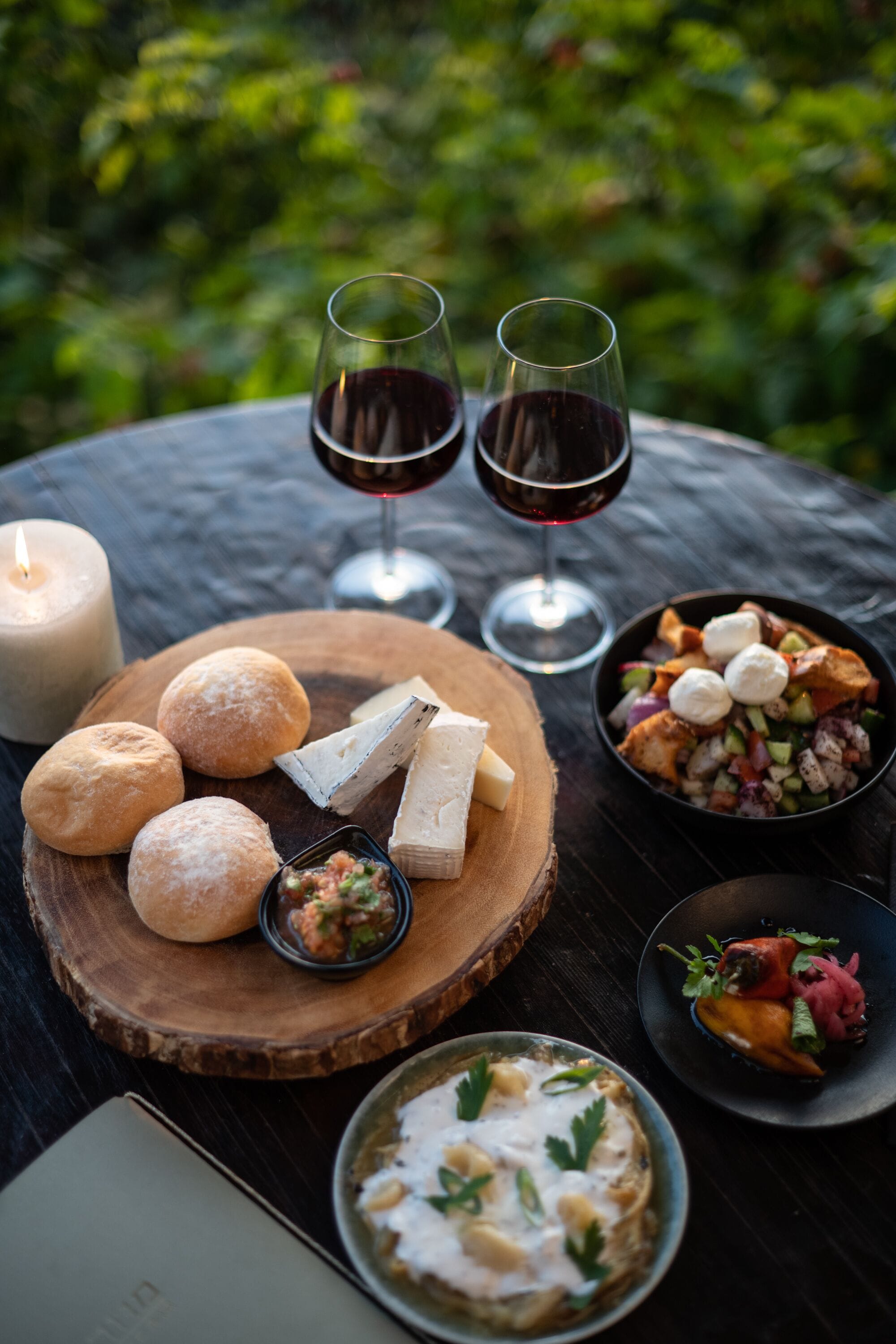 Una mesa dispuesta con varios quesos, pan, un bol de ensalada y dos copas de vino tinto, con una vela y un libro cerca.