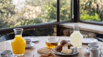 A breakfast table with various food items and drinks, a view of trees through large windows, and a stone wall in the background.