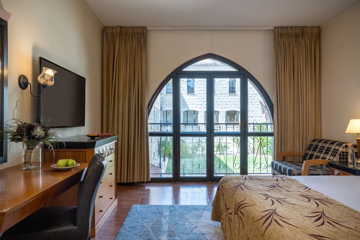 A hotel room with a large arched window, a desk with a vase of flowers, a bed with a patterned bedspread, and a view of a building outside.