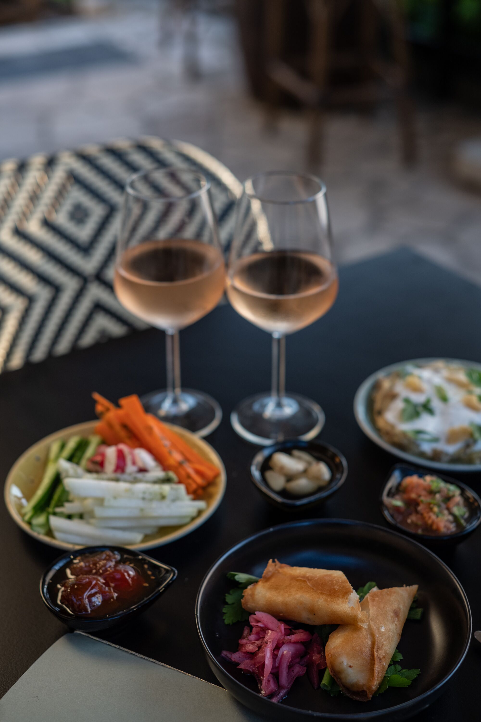 Una mesa con comida y bebidas, incluyendo un plato de rollitos de primavera, un bol de verduras y dos copas de vino.