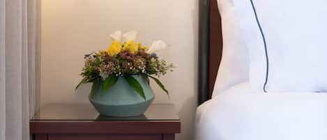 Un lit bien fait, avec une literie blanche et un coussin décoratif, une petite table de chevet en bois sur laquelle est posé un vase de fleurs, et un luminaire fixé au mur.