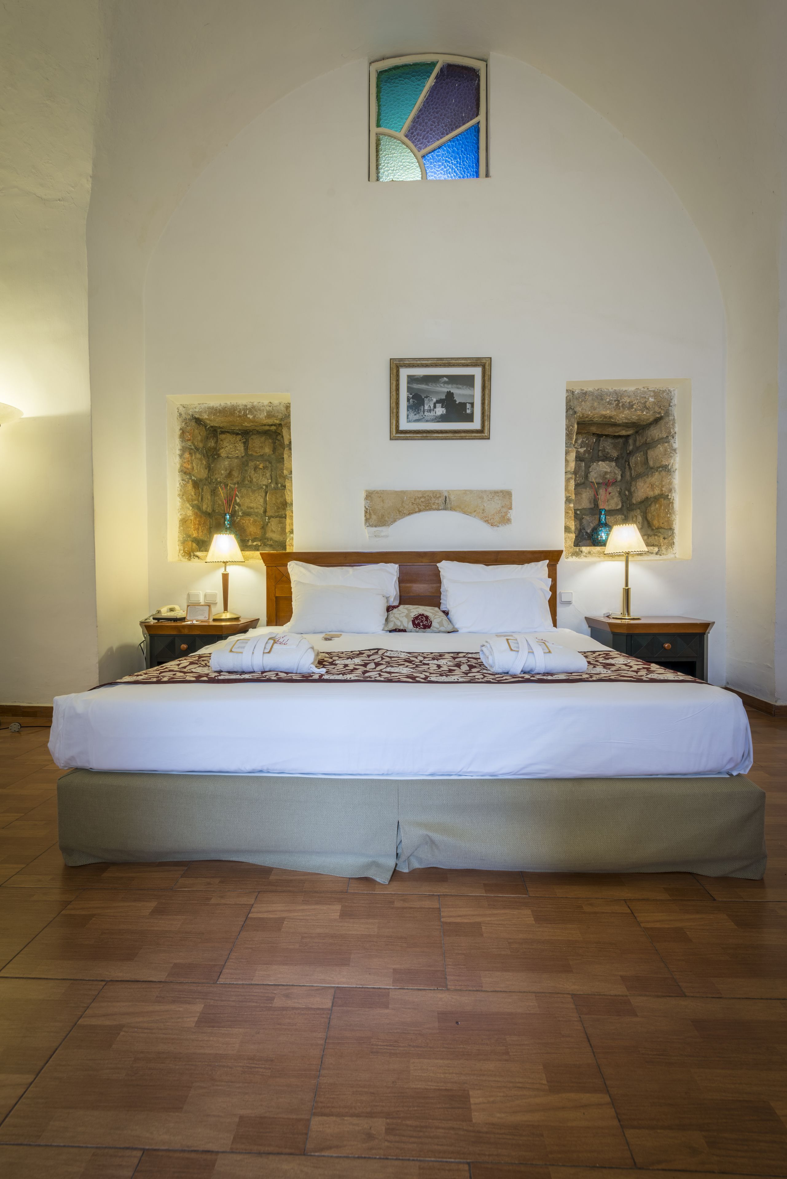 A neatly made bed with white linens, two bedside tables with lamps, and a stone wall with a stained glass window above.