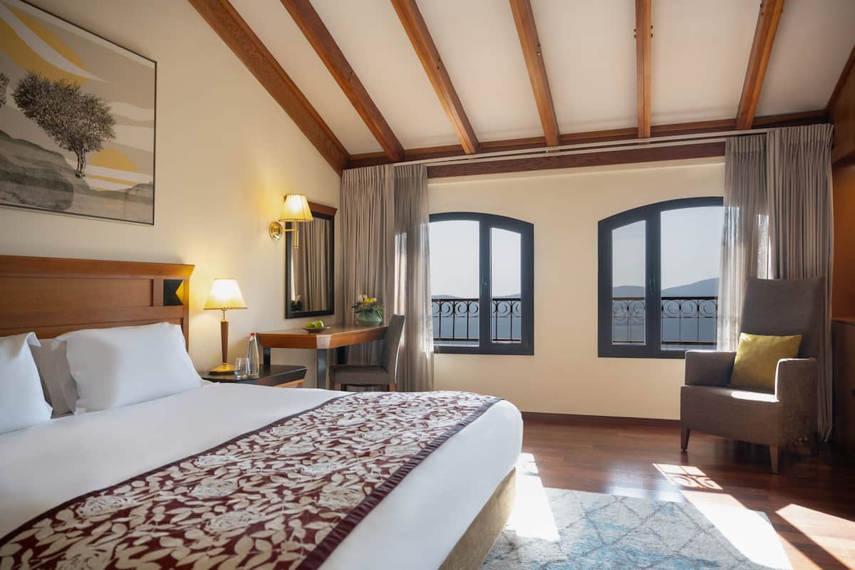 A hotel room with a large bed, a desk with a lamp, a chair, and a view of the outside through two windows.