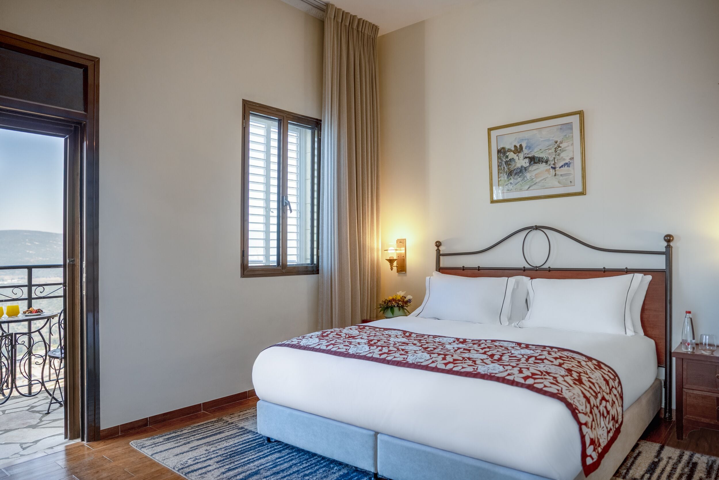 Una habitación de hotel con cama, ventana con cortinas, un cuadro en la pared y un balcón con mesa y sillas.