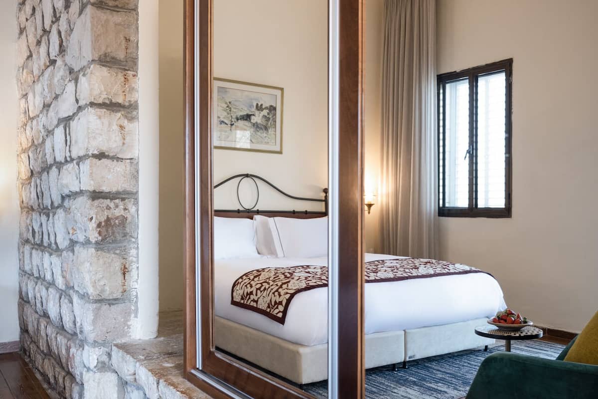 A bedroom with a stone wall, a large mirror, a bed with a white and patterned bedspread, a small table with a bowl of fruit, and a green chair.