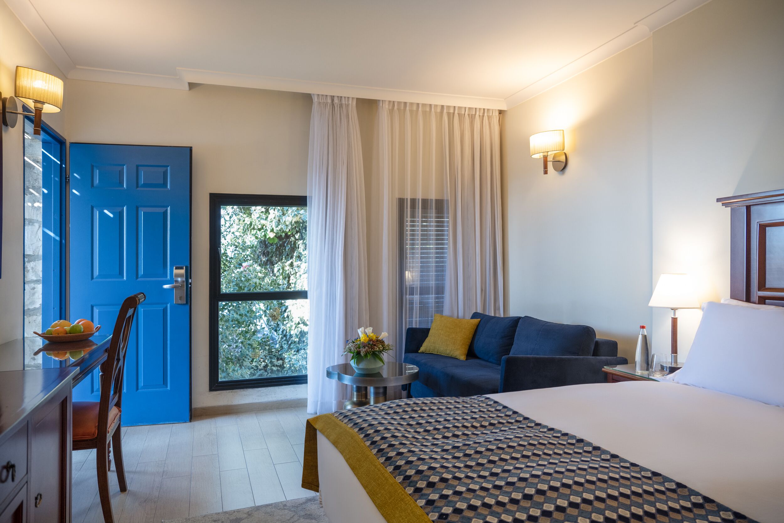 Una habitación de hotel con cama, una puerta azul, una mesa de comedor con sillas, un sofá y una ventana con cortinas.