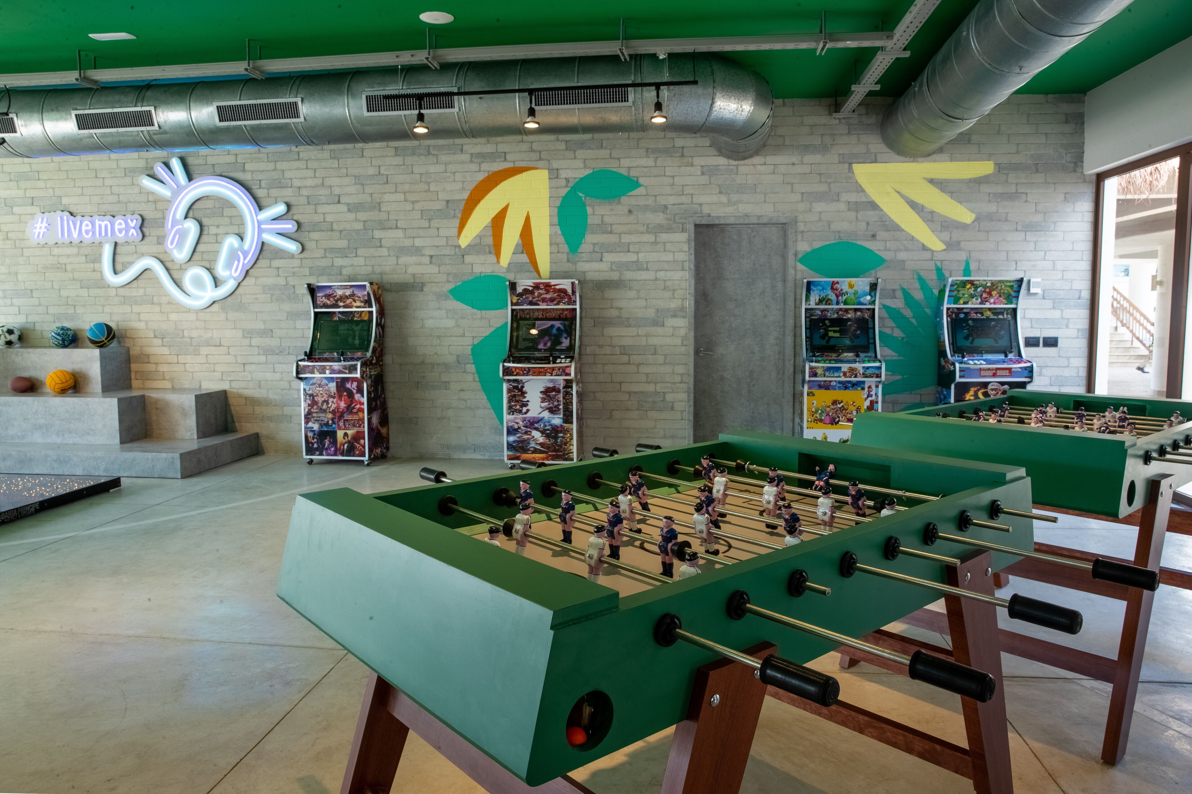 A room with two foosball tables, a neon sign, and arcade games.