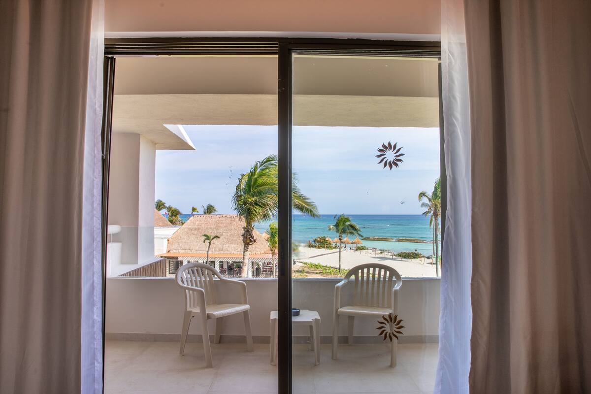 Ein Balkon mit zwei weißen Stühlen und einem kleinen Tisch, der Blick auf einen Strand mit Palmen und ein Gebäude mit Strohdach bietet.