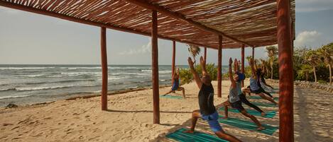 Clase de yoga en la playa bajo una pérgola de madera con vistas al océano.