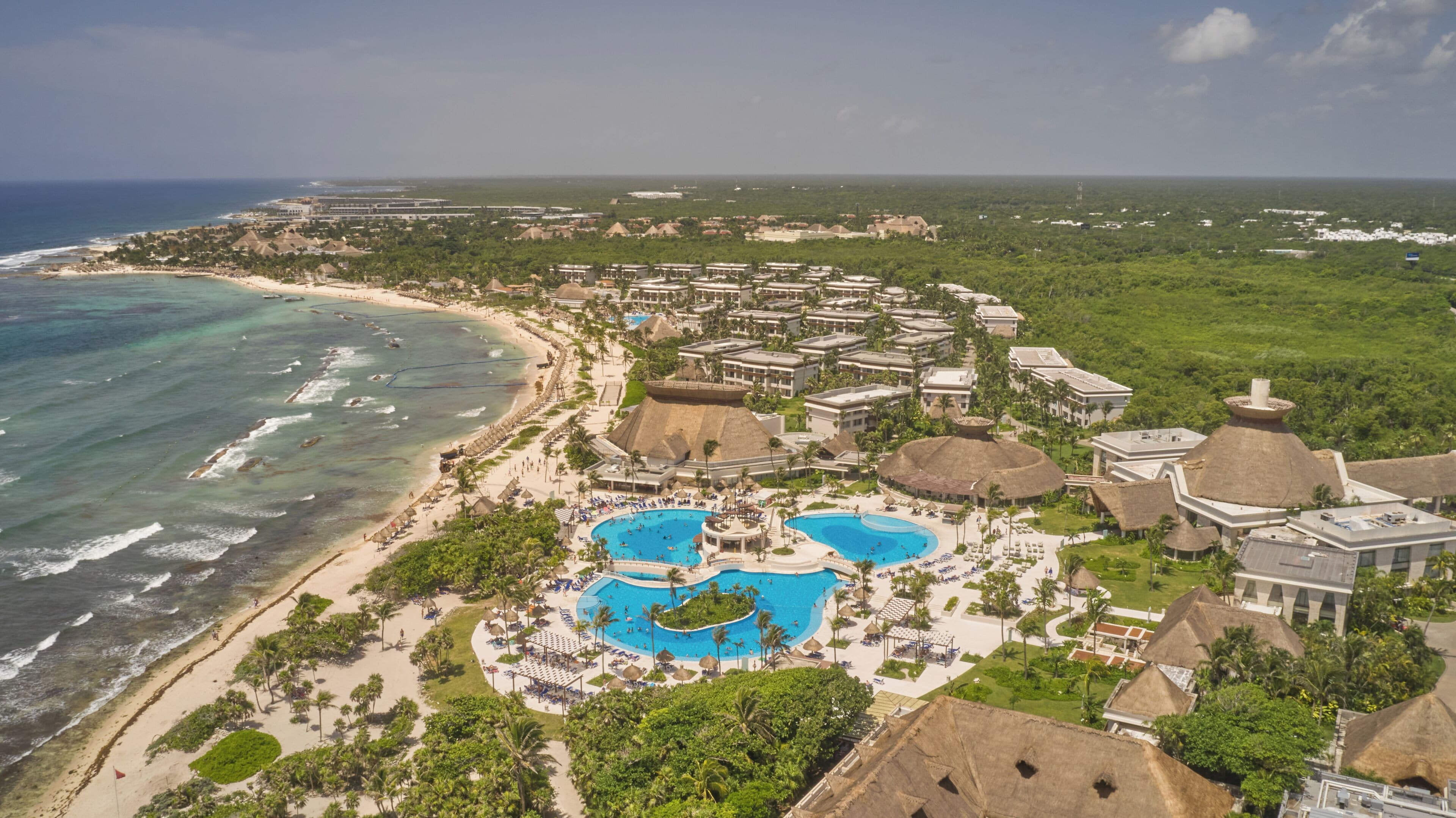 Un resort con una amplia piscina central, acceso a la playa y edificios de techo de paja.