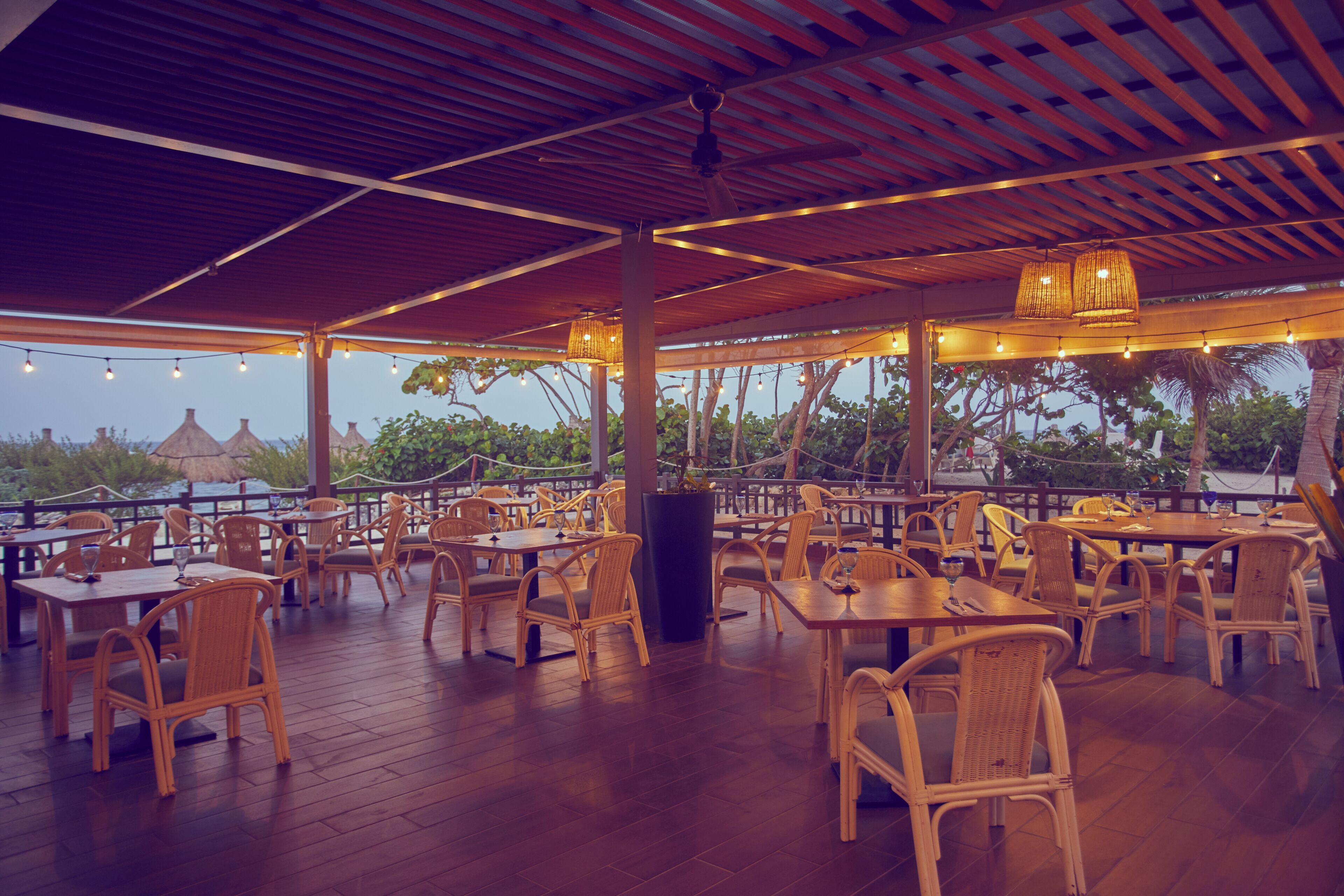 Un área de comedor al aire libre con mesas y sillas de madera, techo cubierto y luces colgantes.