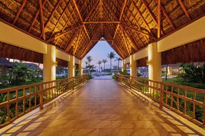 A covered walkway with a thatched roof, palm trees, and a view of a resort area. - Bahia Principe Explore Tulum - All Inclusive (Akumal)