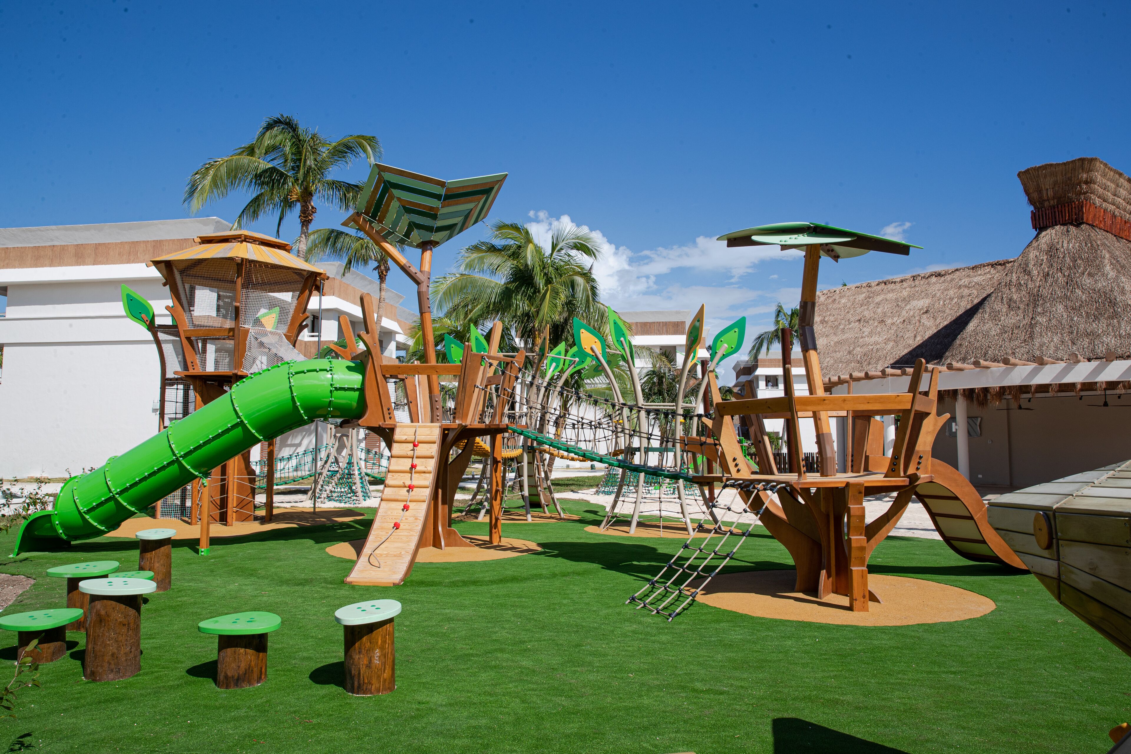 Un parque infantil con un tobogán verde, estructuras para escalar y mesas sobre césped artificial.
