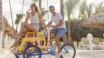 Un couple se promène sur une plage, poussant un chariot sur lequel on peut lire le mot « BON », avec un palmier en arrière-plan.