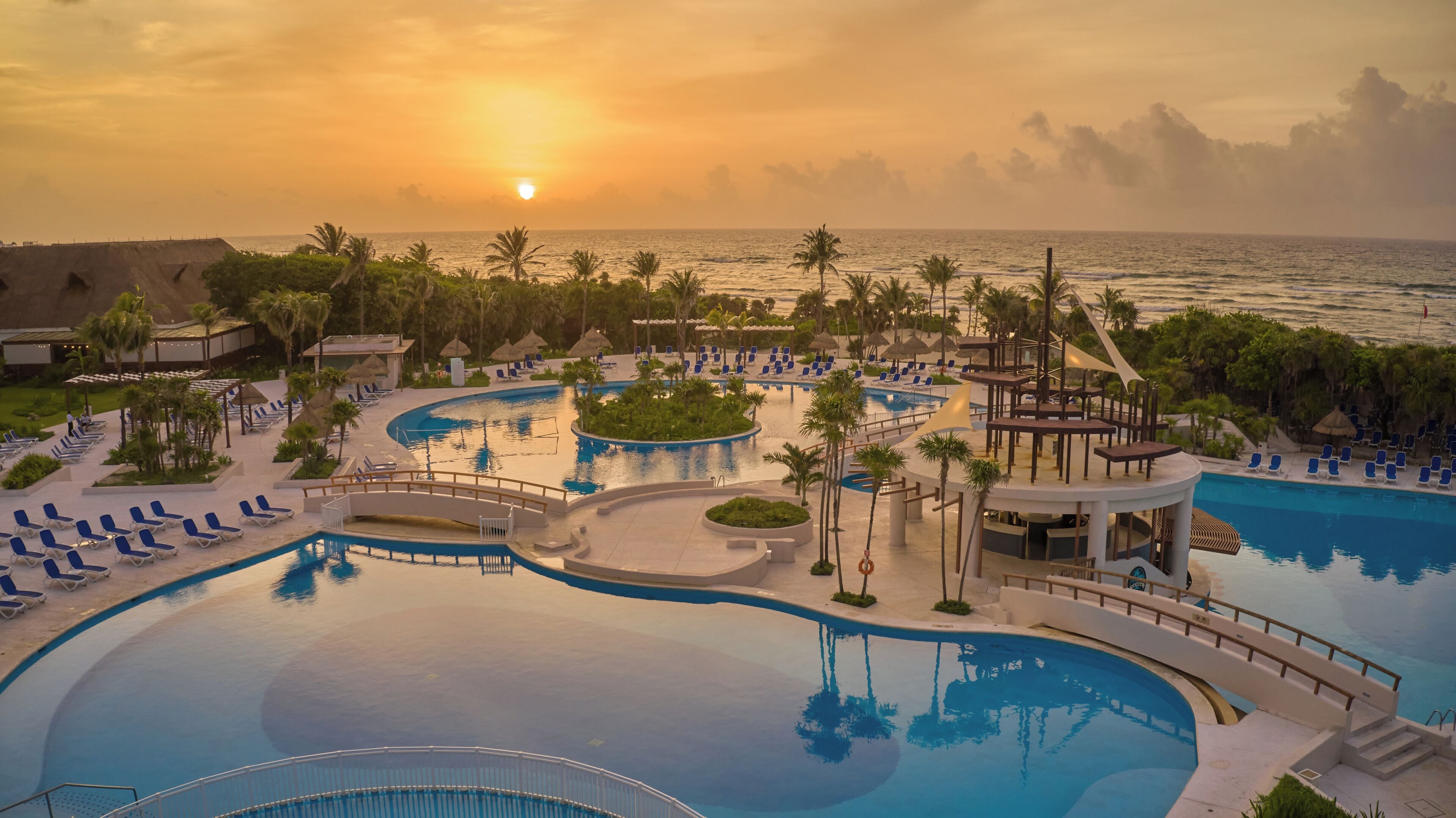 Un resort con una piscina grande, tumbonas y vista al océano al atardecer.