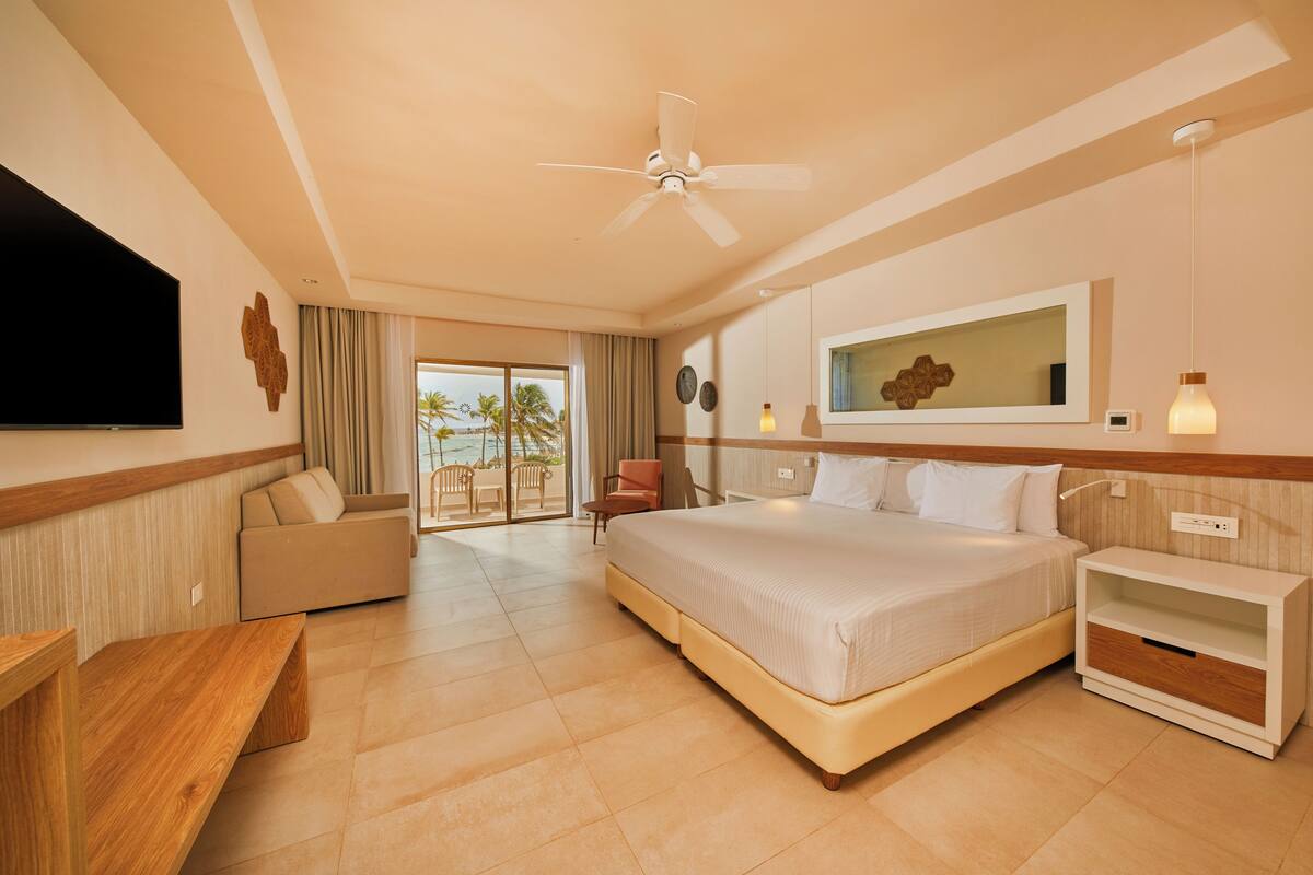 A hotel room with a large bed, a TV, a ceiling fan, a sofa, a desk, and a balcony with a view of palm trees.