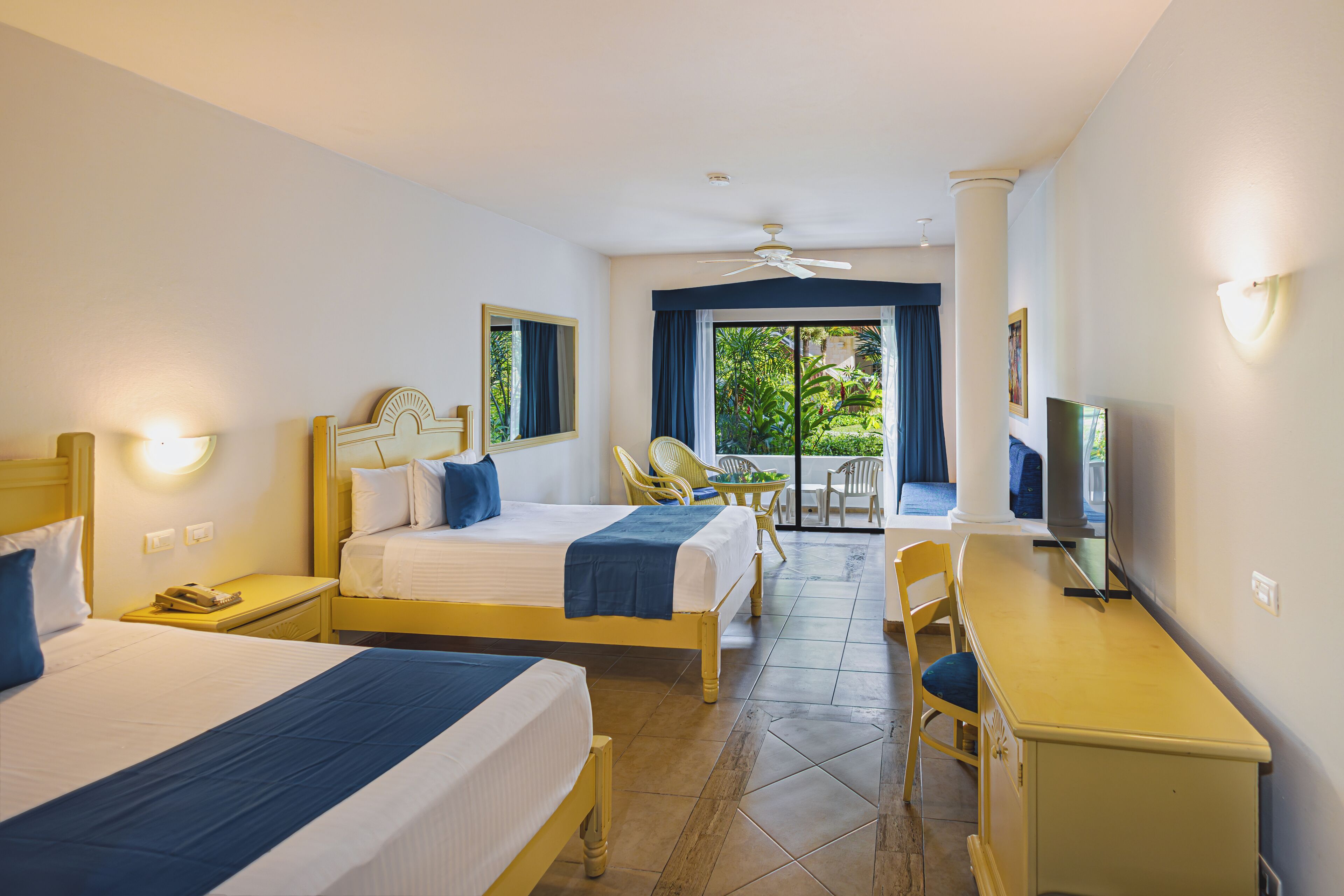 Quarto de hotel com duas camas, uma escrivaninha, televisão e varanda com vista para o verde.