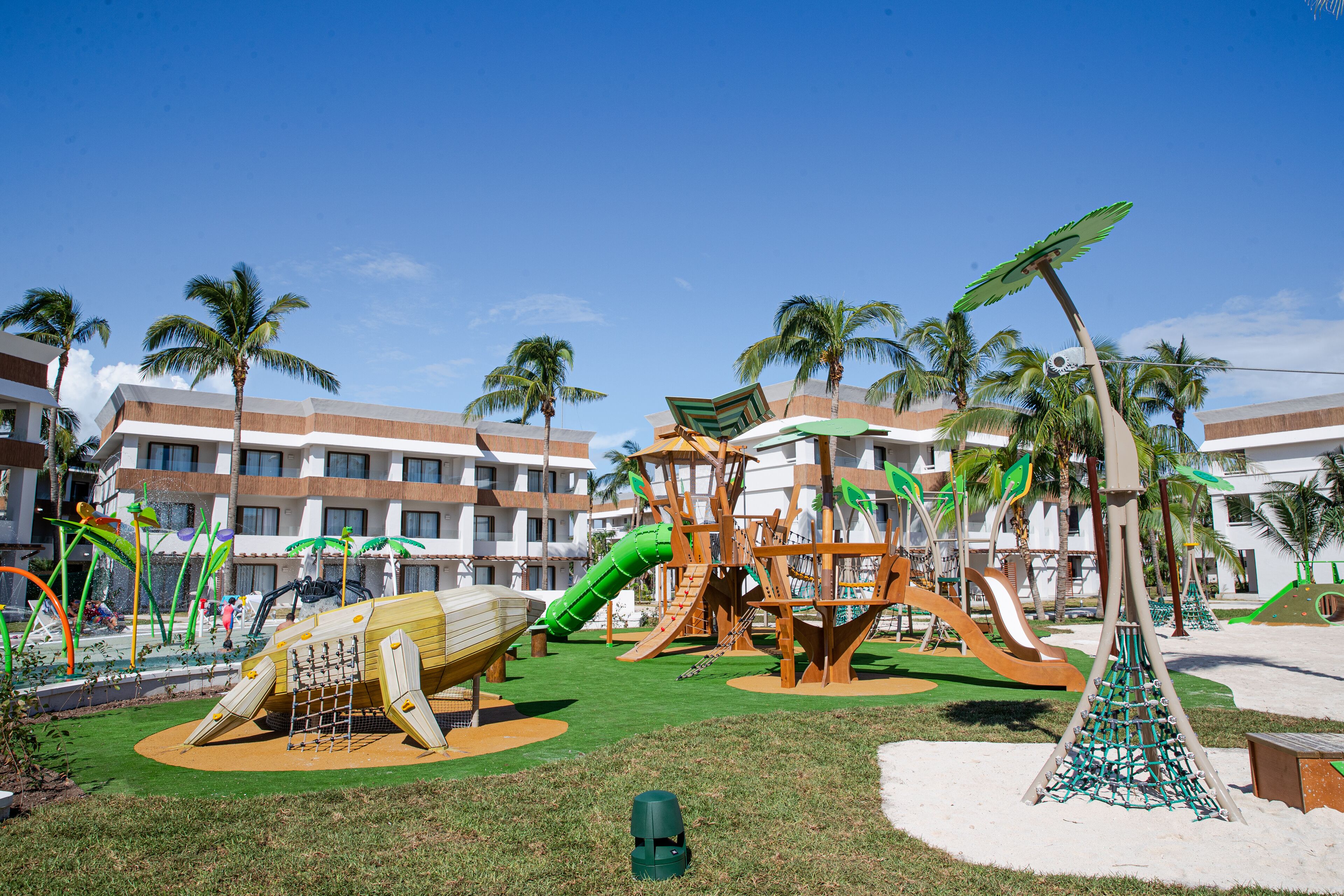 Un parque infantil con toboganes y estructuras para escalar, rodeado de palmeras y un edificio con balcones.
