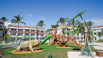Un parque infantil con toboganes y estructuras para escalar, rodeado de palmeras y un edificio con balcones.