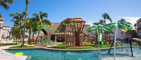 A pool area with a water slide, palm trees, and a building with a thatched roof.