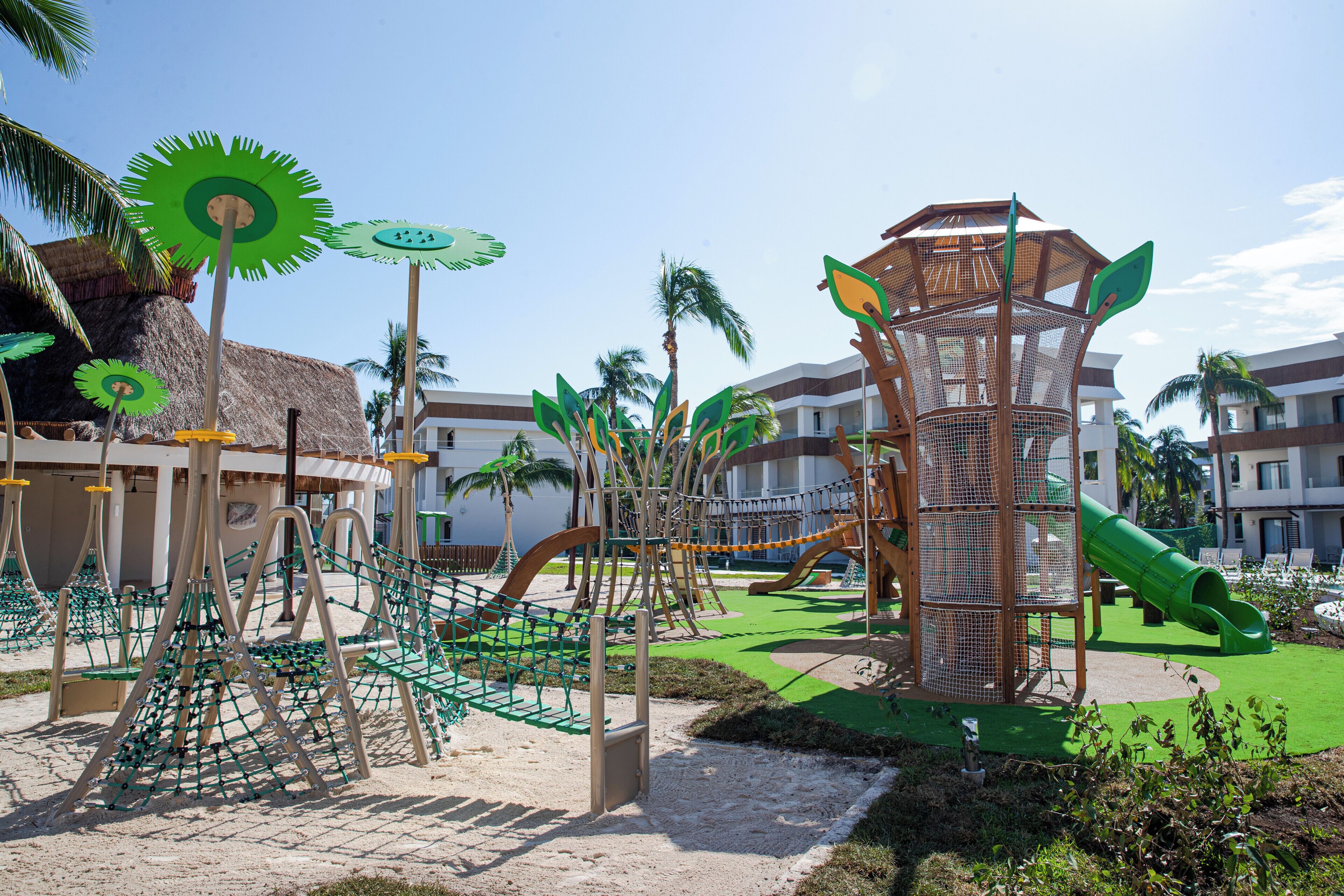 A playground with climbing structures and slides, surrounded by palm trees and buildings.