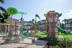 Un parque infantil con estructuras para escalar y toboganes, rodeado de palmeras y edificios.