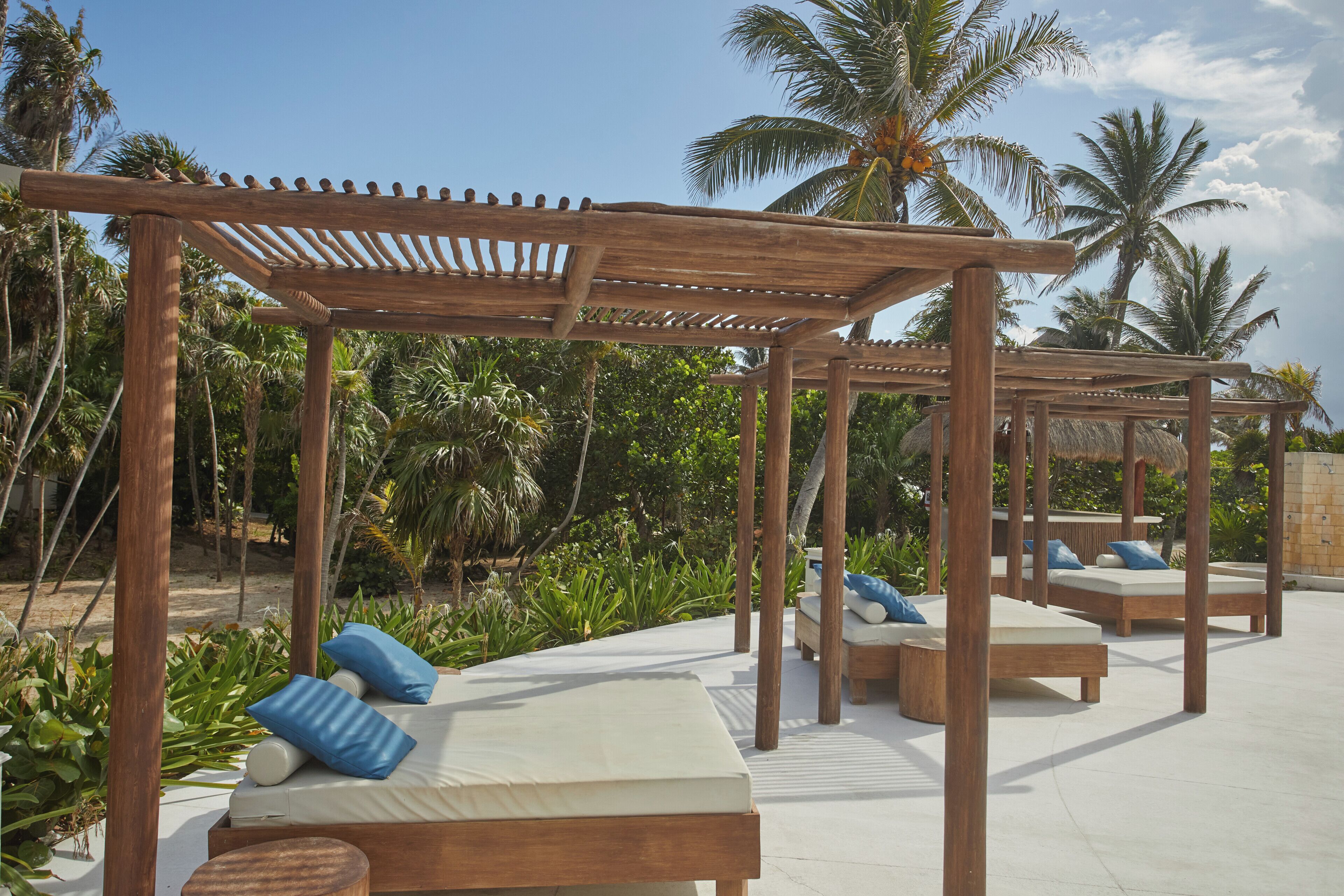 A wooden pergola structure with lounge beds and blue pillows, surrounded by palm trees and greenery.