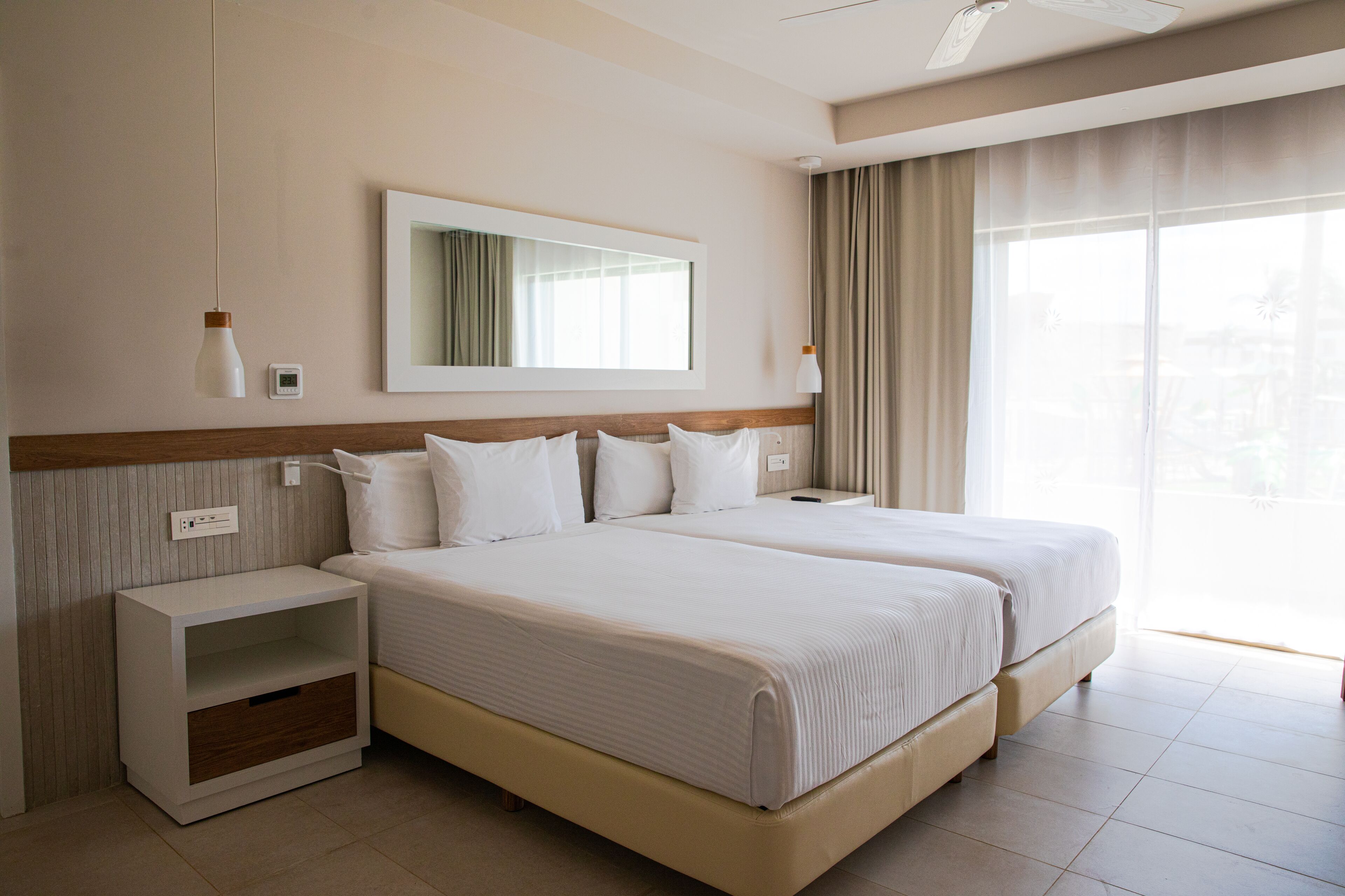 A hotel room with two beds, a nightstand, a mirror, and a large window with curtains.