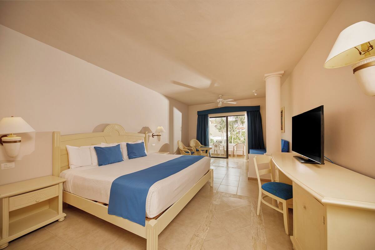 A hotel room with a bed, desk, TV, and a balcony with chairs.