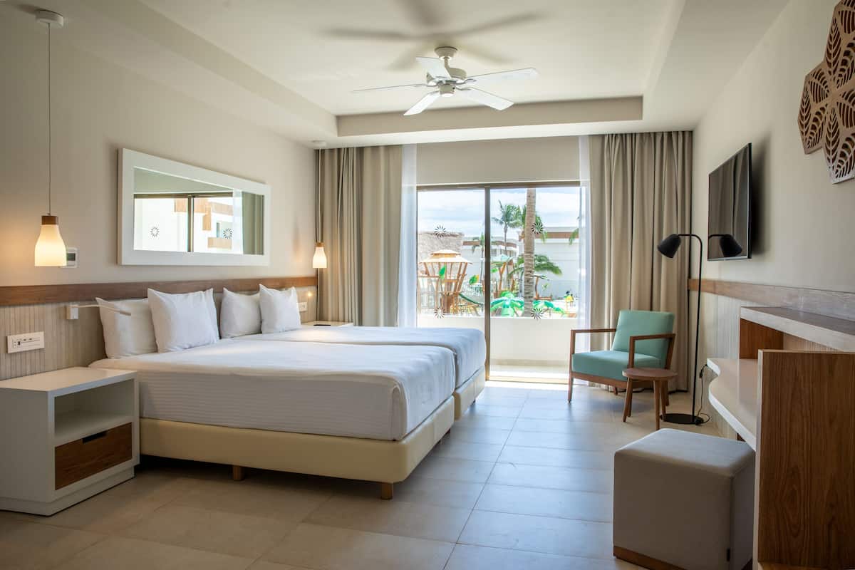 Ein Hotelzimmer mit einem großen Bett, einem Schreibtisch, einem Sessel, einem Fernseher und einem Balkon mit Blick auf Palmen und einen Pool.