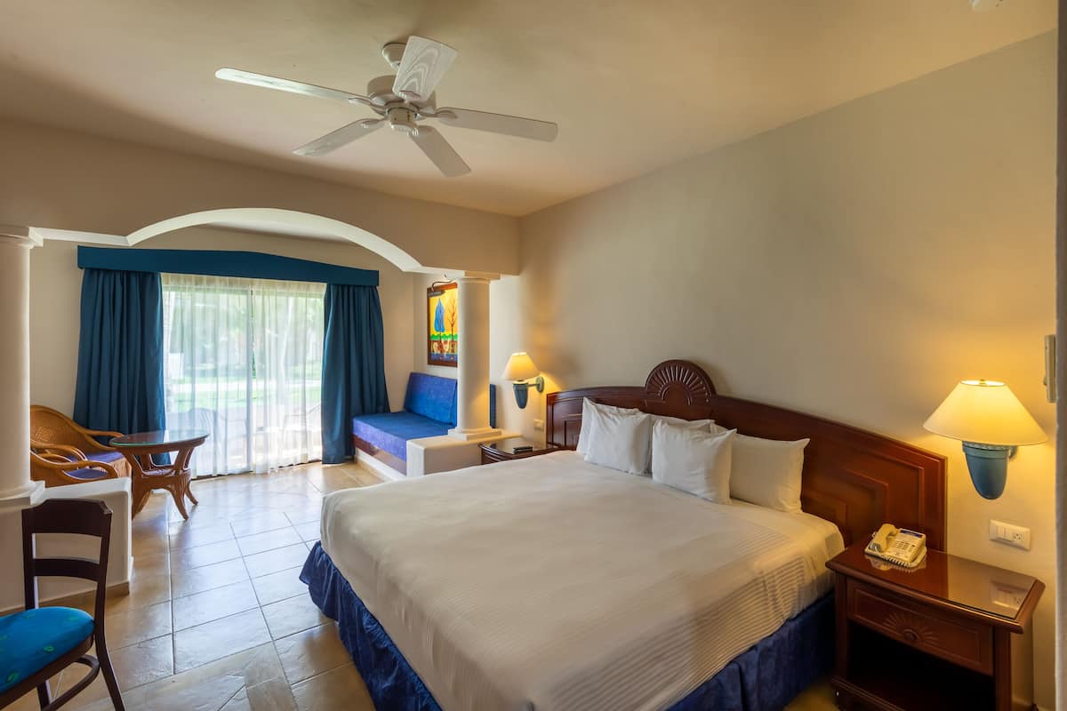 Ein Hotelzimmer mit einem großen Bett, einem Schreibtisch, einem Sessel, einem Deckenventilator und einem Fenster mit Vorhängen.