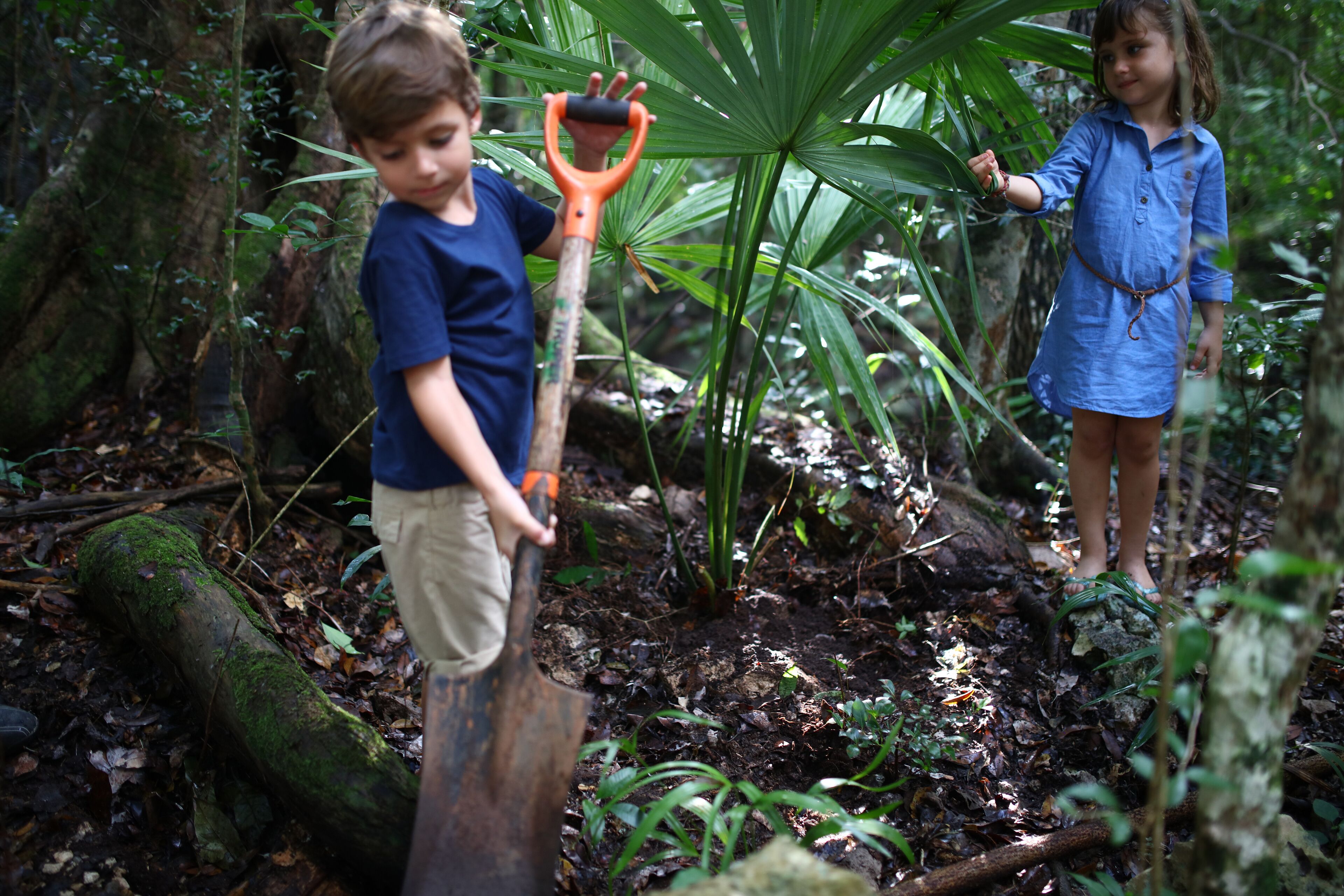 Two children in a forested area, one holding a shovel.
