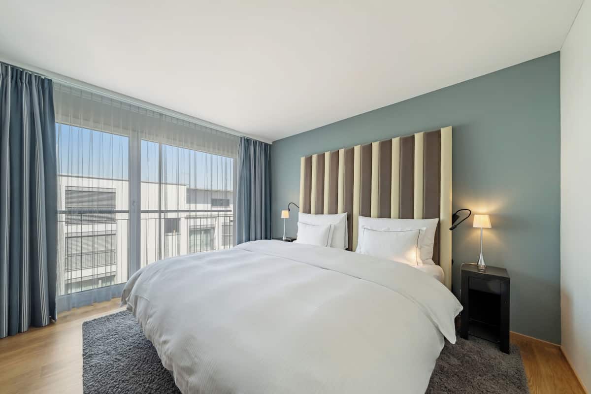 Una habitación de hotel moderna con una cama grande, dos mesitas de noche con lámparas, vistas a la ciudad a través de amplios ventanales y una alfombra gris.