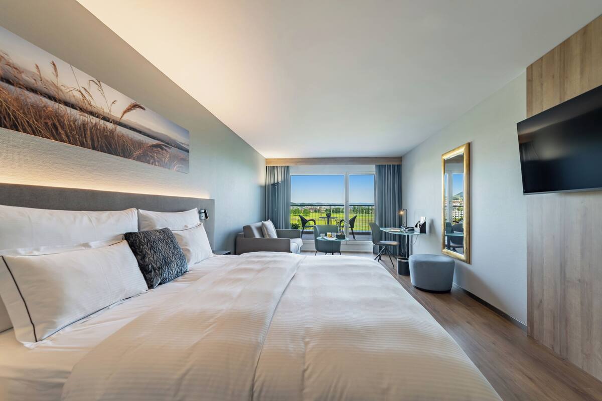 Una habitación de hotel moderna con una cama grande, un televisor de pantalla plana, una zona de comedor con vistas y un cuadro en la pared.