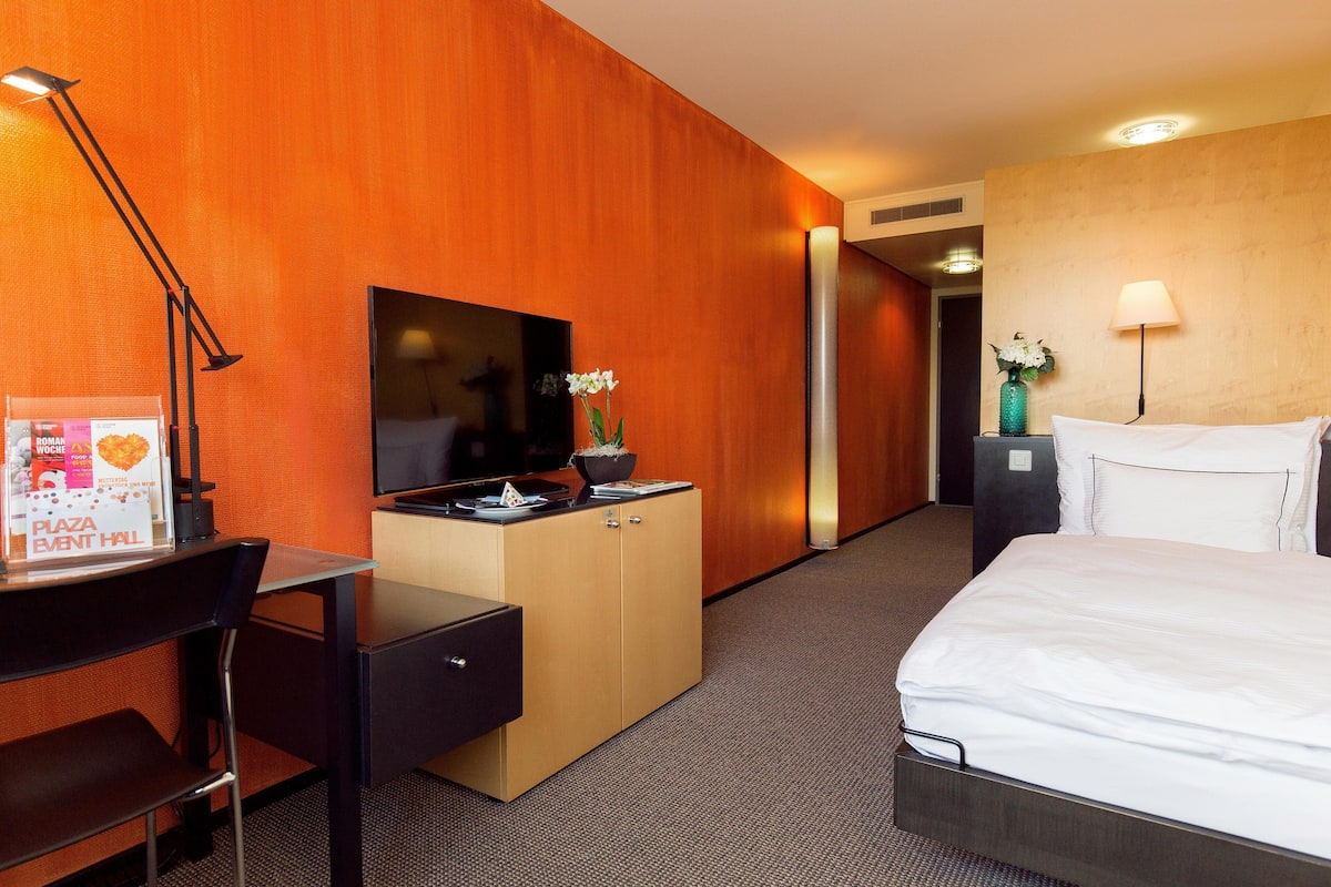 Habitación de hotel con cama, escritorio con silla, televisor, minibar y lámpara de pared.