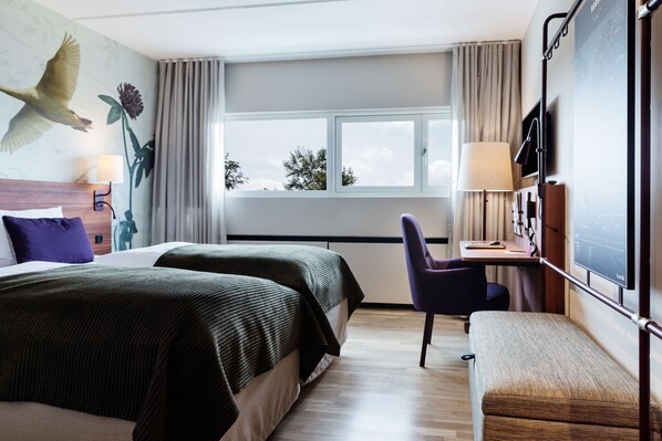 A hotel room with two beds, a desk, a chair, a TV, and a window with a view of trees.