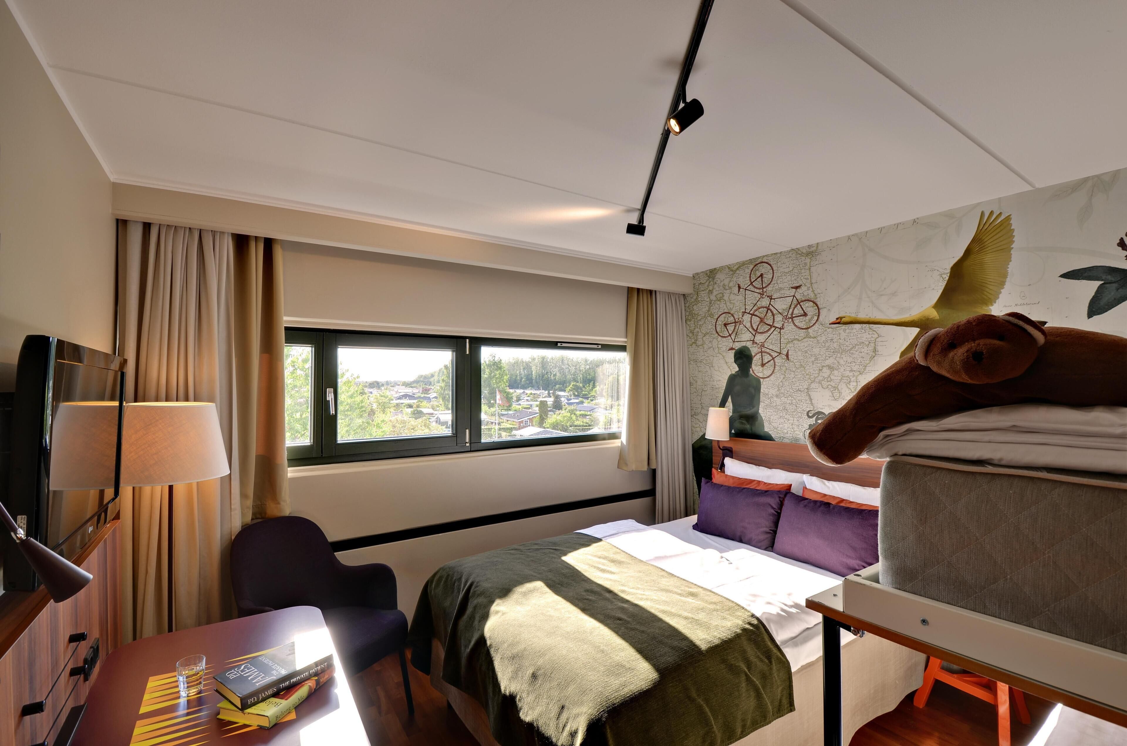 A hotel room with a bed, a desk with a lamp, a chair, a window with a view, and a wall with a mural.
