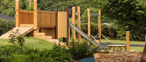 A wooden play structure with a slide and steps, surrounded by greenery and mulch.