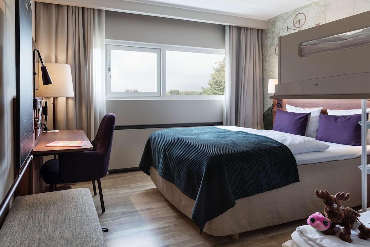 A hotel room with a bed, a desk, a chair, a lamp, and a window.