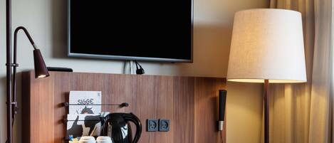 A hotel room with a wooden desk, a flat-screen TV mounted on the wall, a lamp, a coffee maker, and a laptop.