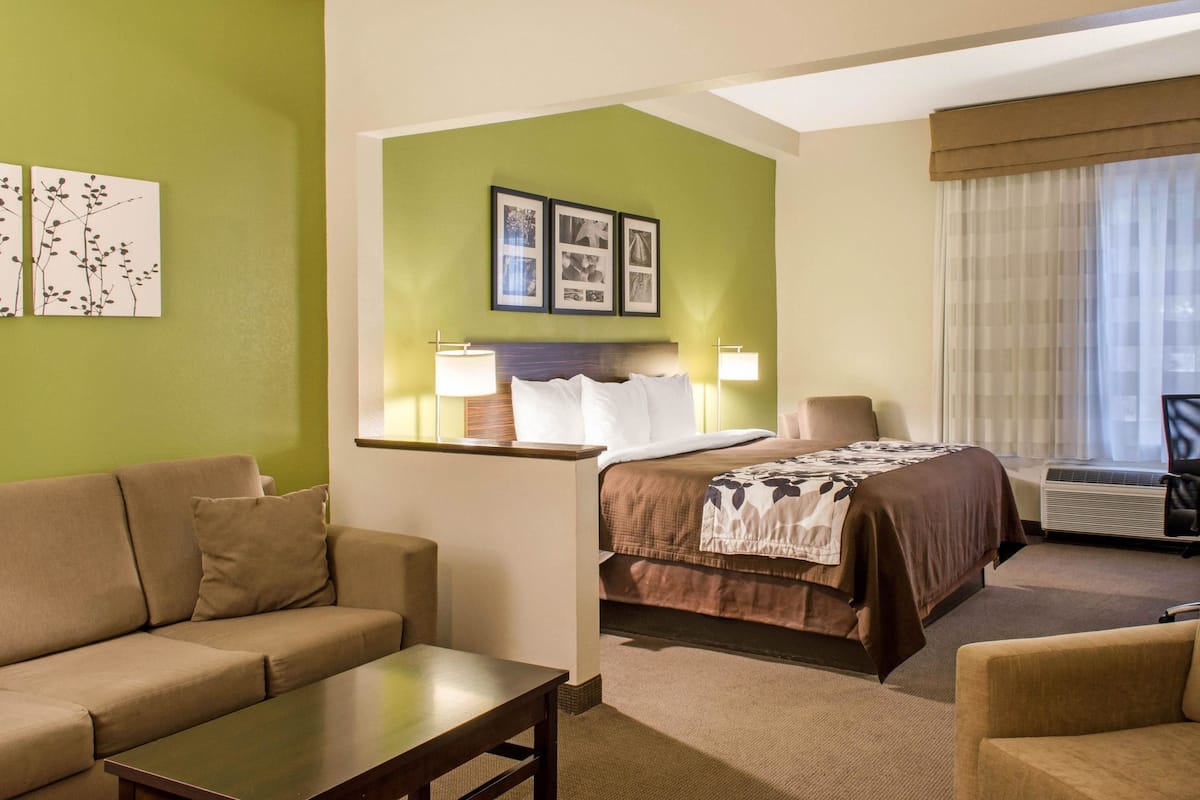 A hotel room with a bed, a sofa, a small table, and a chair. There are framed pictures on the wall and a window with curtains.