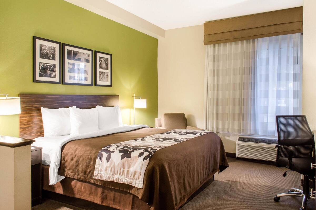 A hotel room with a large bed, a desk with a lamp, a chair, and a window with curtains.