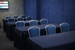 Una sala conferenze con file di tavoli e sedie, uno schermo per proiettore e un muro blu.