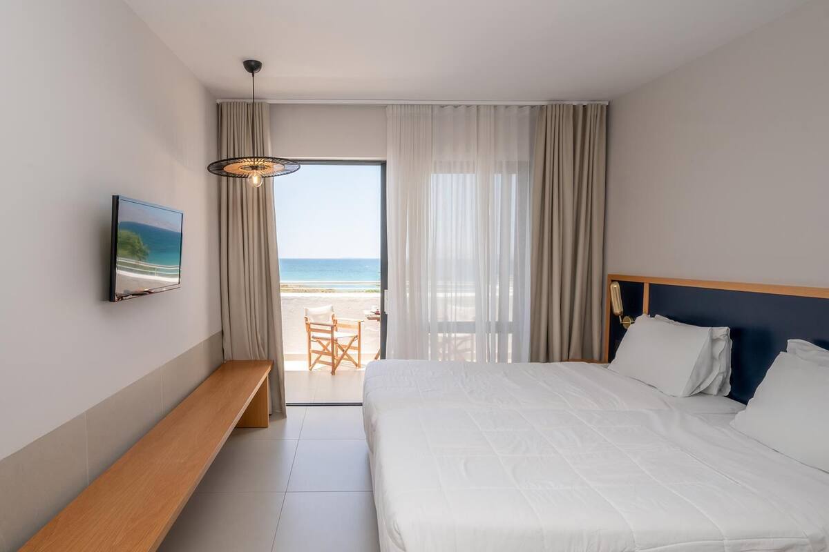 Habitación de hotel con cama, un banco, televisor y vista a la playa.
