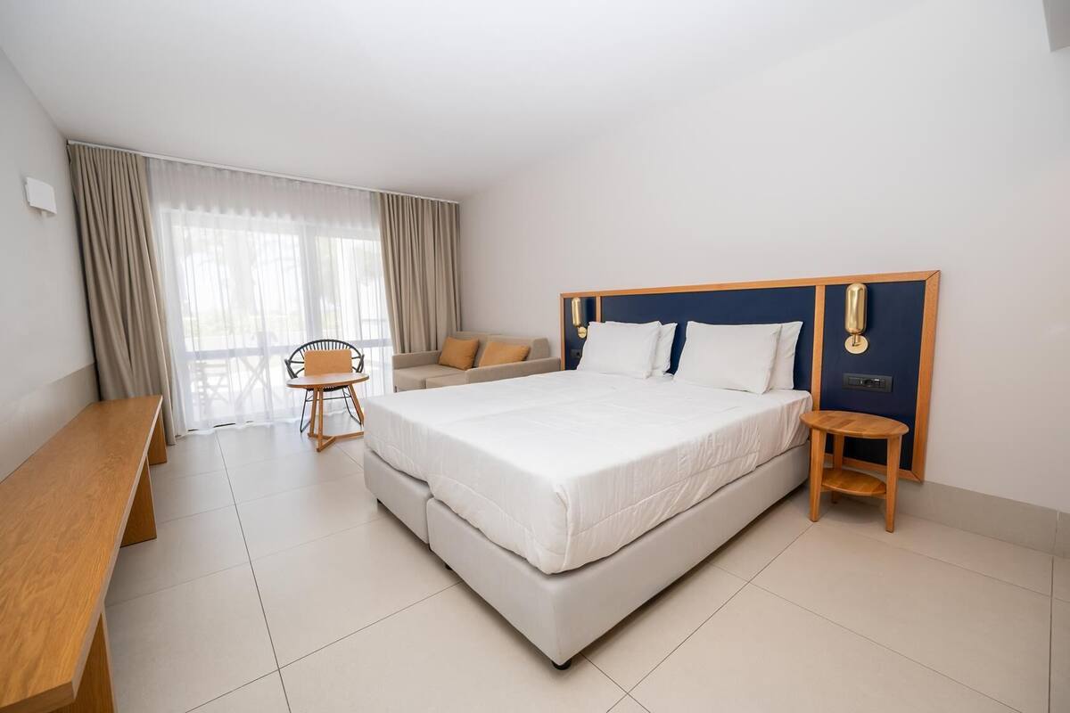 Una habitación de hotel moderna con una cama grande, un banco de madera, una mesita pequeña y un sofá.