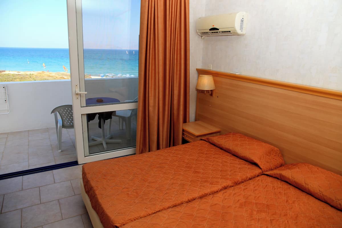 Un cuarto de hotel con cama, mesita de noche, lámpara, ventana con cortinas y vista a la playa y al océano.
