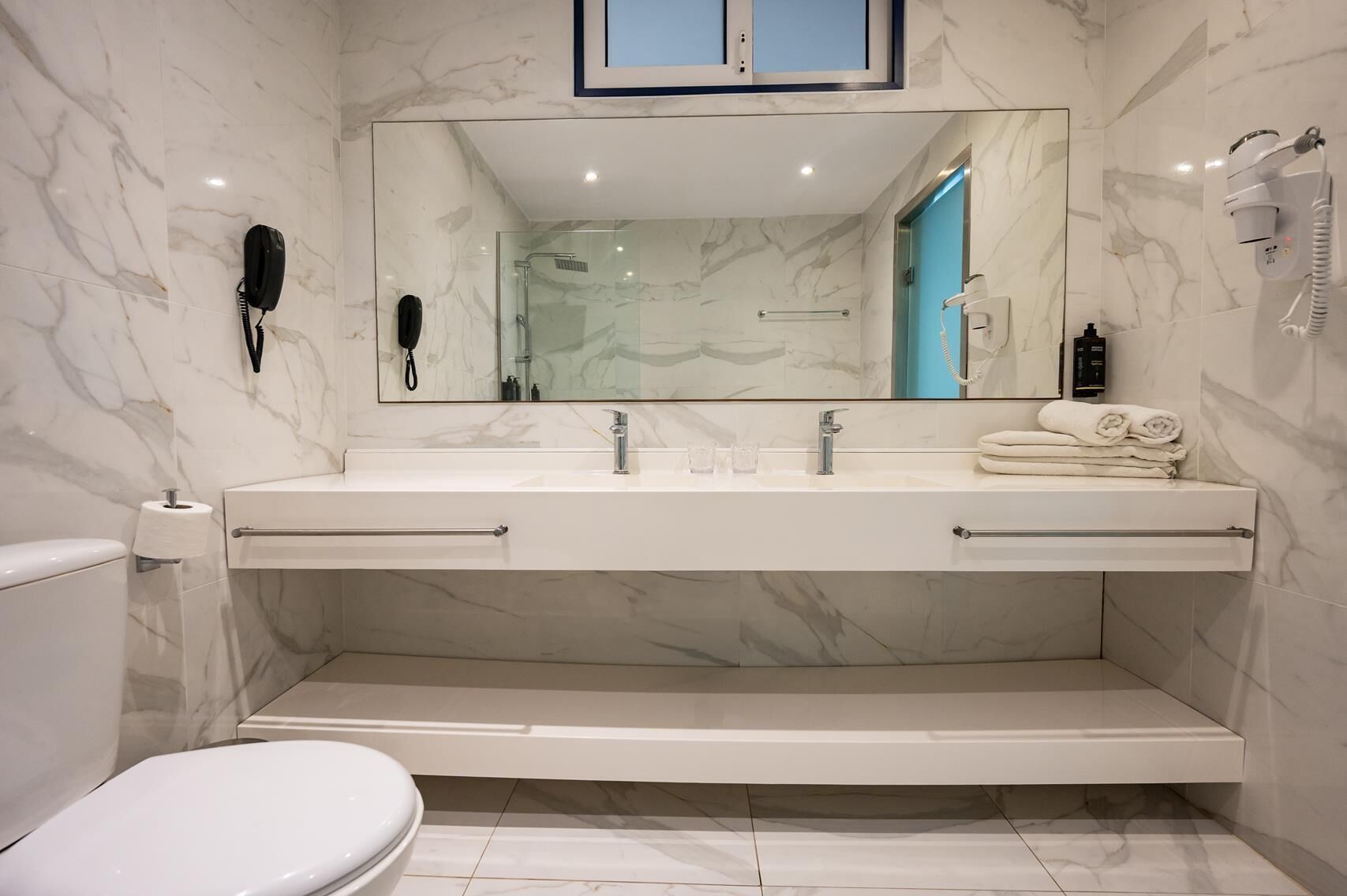 Un baño moderno con suelo de mármol, un espejo grande, lavabo doble, inodoro y zona de ducha.